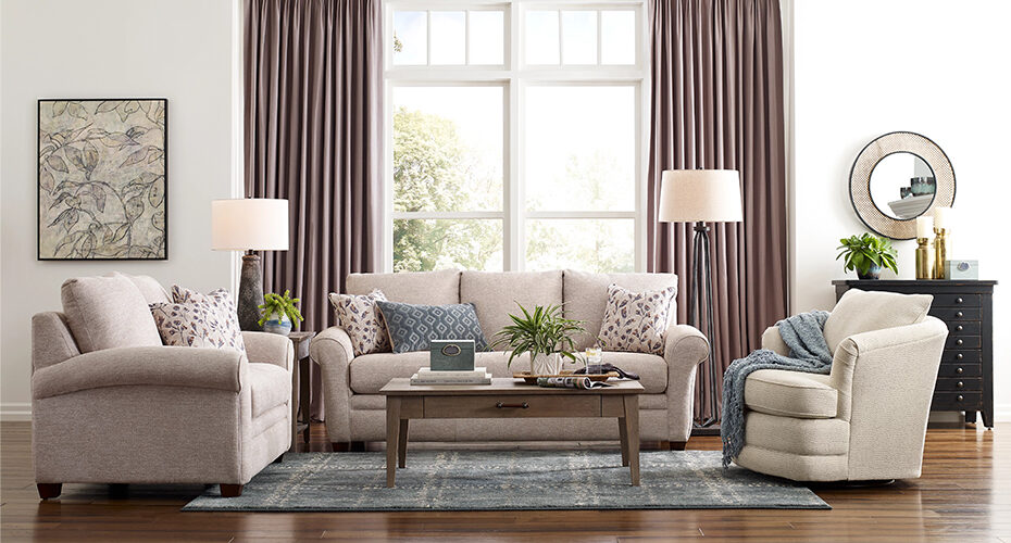 A cozy living room features a comfortable setup with a gray sofa, an armchair, and a round swivel chair arranged around a wooden coffee table. Two floor lamps, a round mirror, and stylish curtains frame a large window, complemented by a framed artwork on the wall.