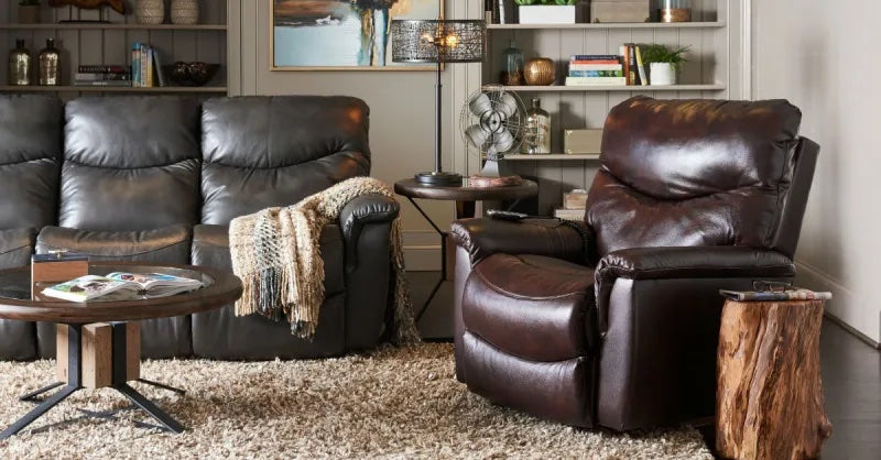A cozy living room with a plush beige rug, a dark brown leather recliner, a black leather loveseat with a knitted throw blanket, and a wooden coffee table with magazines. The room features bookshelves, decorative items, and a floor lamp with a fan on a side table.