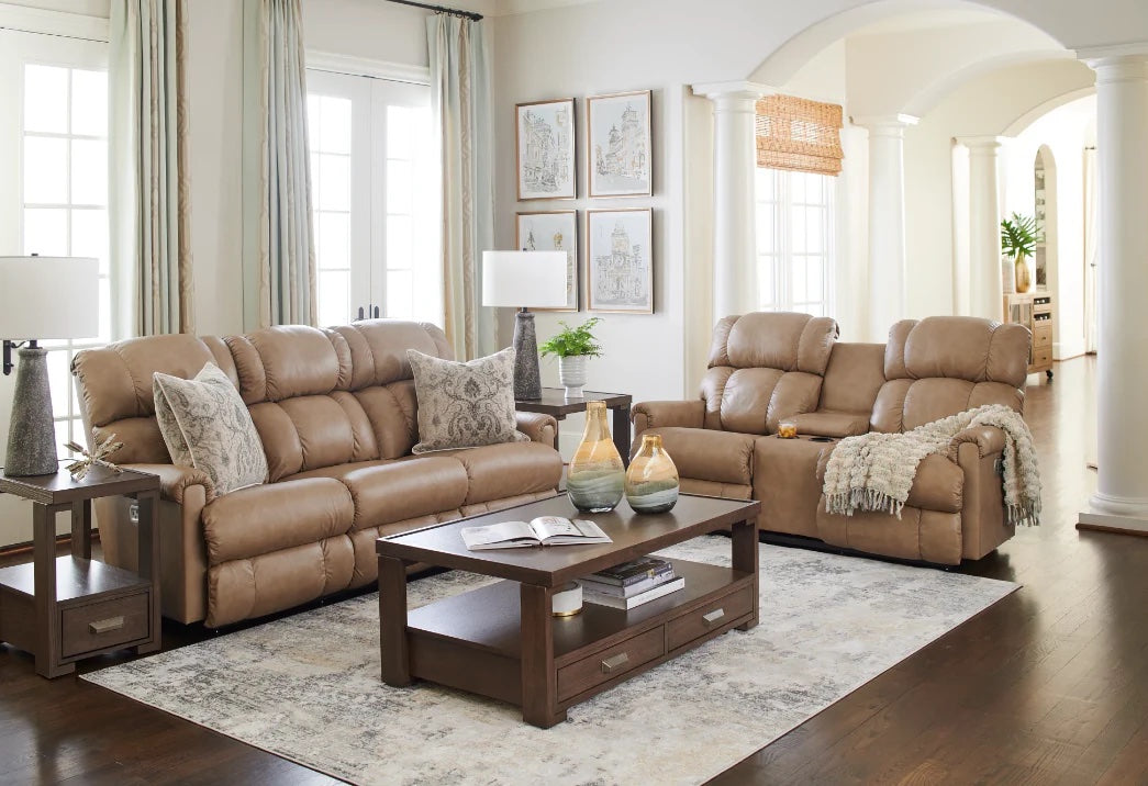 A cozy living room with a beige leather sofa and matching loveseat, both adorned with decorative pillows and a throw blanket. A wooden coffee table holds decorative vases and books, and side tables with lamps flank the seating. Large windows and artwork decorate the space.