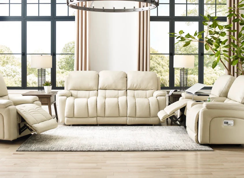A cozy living room features a set of cream-colored reclining sofas and armchairs. A large window with beige curtains allows natural light to flood the space. Two side tables with lamps and a green plant add decorative elements. A neutral area rug covers the hardwood floor.