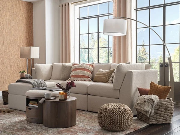 A cozy living room features a beige sectional sofa adorned with throw pillows, a beige pouf, a round wooden coffee table with decor, and a basket with blankets. Large windows with beige curtains reveal a scenic mountain view. Floor lamp provides soft lighting.