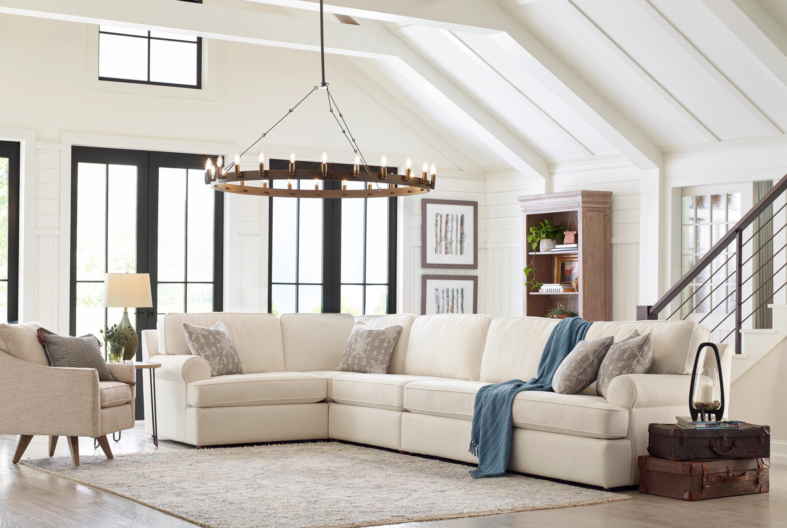 A spacious living room features a large, cream-colored sectional sofa adorned with patterned cushions. A modern chandelier hangs from the high, vaulted ceiling. A cozy armchair sits nearby, and a bookshelf stands against the wall. Light floods through large windows.