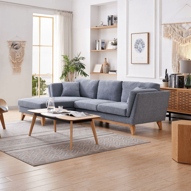 A modern living room featuring the ValMinimal Sectional, a gray upholstered sectional sofa with wooden legs. In the center, there is a wooden coffee table adorned with a glass pitcher and cups. The room is further enhanced by plants, wall art, and shelves. Sunlight streams through a large window.