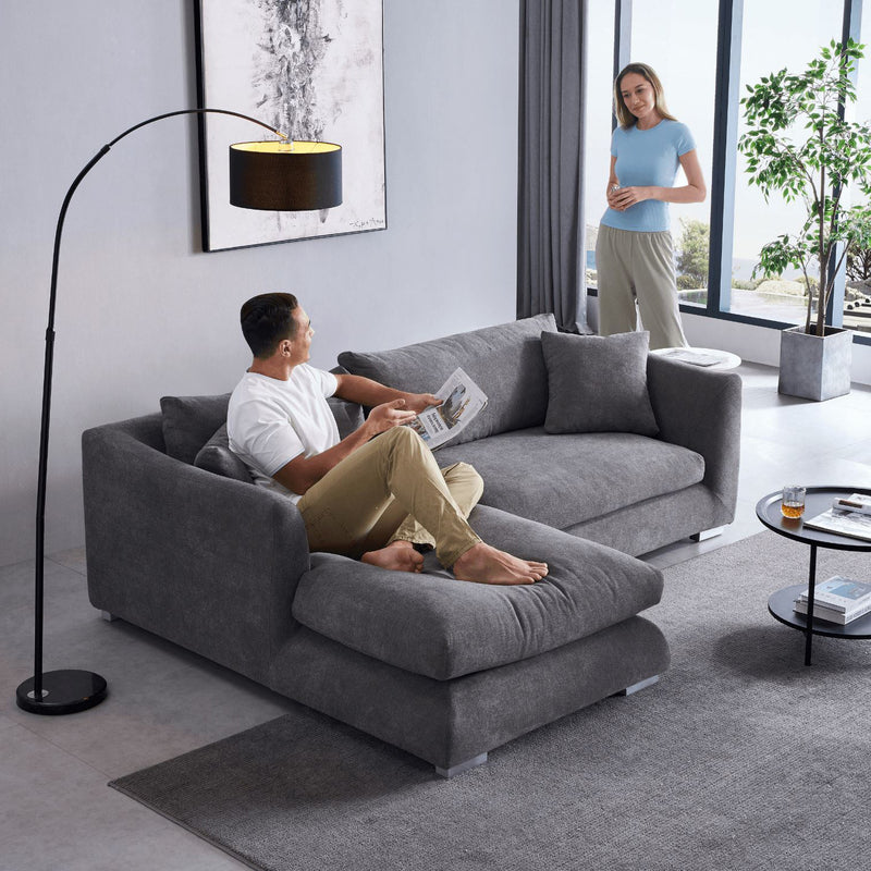 A man sits on the Feathers 88" Left Facing Sectional, reading a newspaper, as a woman stands nearby with a smile. The room's modern design includes a floor lamp, a round coffee table, and large windows that offer views of trees and the sky.