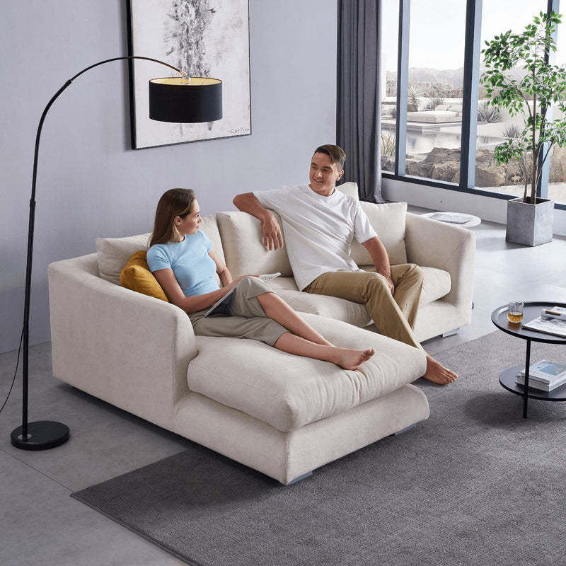 A couple relaxes on the Feathers 88" Left Facing Sectional in a bright living room. The woman leans back with her legs extended, while the man gestures conversationally. A black floor lamp, abstract artwork, and large windows enhance the space around them.