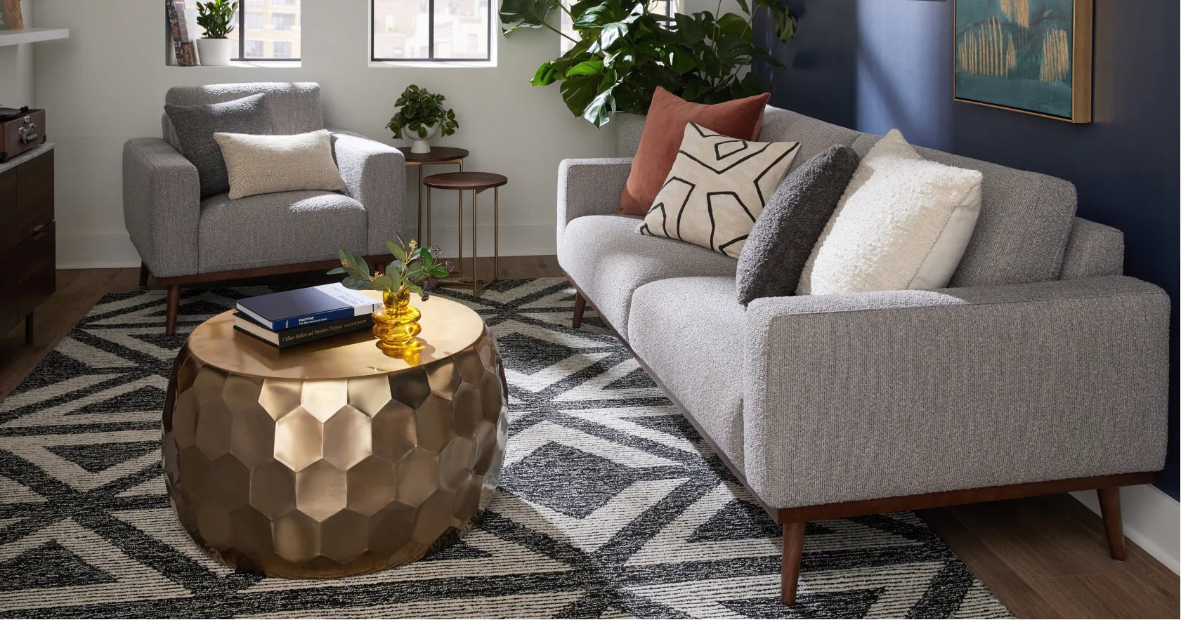A modern living room with a large window showing a garden view. It features a gray sofa, brown leather armchair, wooden coffee table, and patterned rug. Decor includes a wall art of a cactus, woven baskets, a wooden sideboard, and a pendant light.