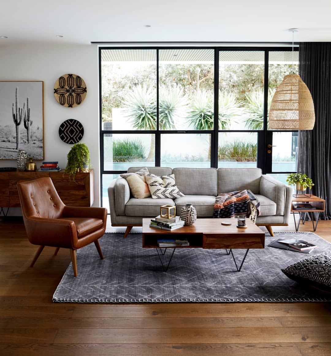 A cozy, light-toned living room features a beige sofa with matching armchair, decorated with blue and gray pillows. A wooden coffee table with storage sits on a patterned rug. A side table with a lamp and decor, along with tall windows and wall art, complete the space.