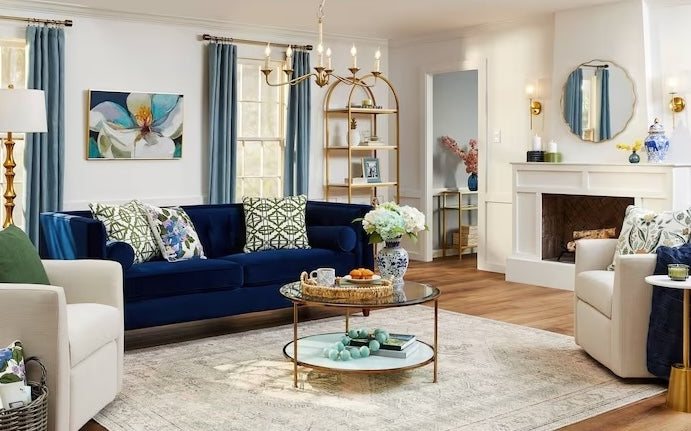 A cozy and elegant living room with a navy-blue velvet sofa adorned with patterned accent pillows. A glass coffee table with floral d√©cor sits atop a light rug. The room features gold accents, a white fireplace, bookshelves with decor items, and large windows with teal curtains.