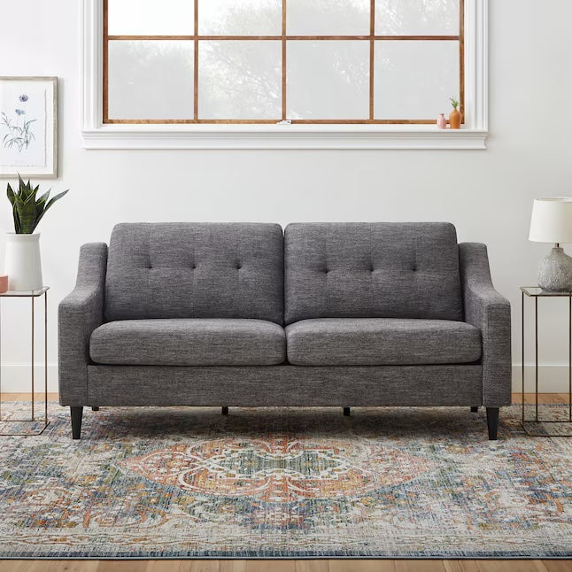 A modern living room features a grey upholstered sofa with button tufting, set against a large window. Two side tables each hold a potted plant and a lamp. The floor is covered with a multicolored patterned rug, and a framed botanical print hangs on the wall.