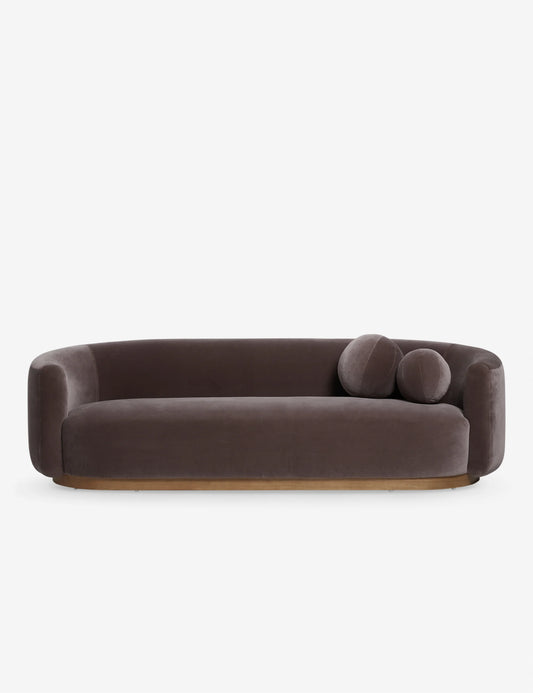 The Lowry Sofa, a contemporary piece with an oval shape and soft rounded edges, comes in a brown hue and includes two matching round cushions. It is elegantly displayed against a plain white background.