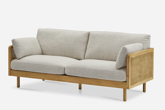 The Lucia Cane Sofa in a whitewash finish showcases a modern design with a wooden frame and light beige cushions, featuring woven rattan sides. Its sleek, minimalist appearance stands out against a plain background.