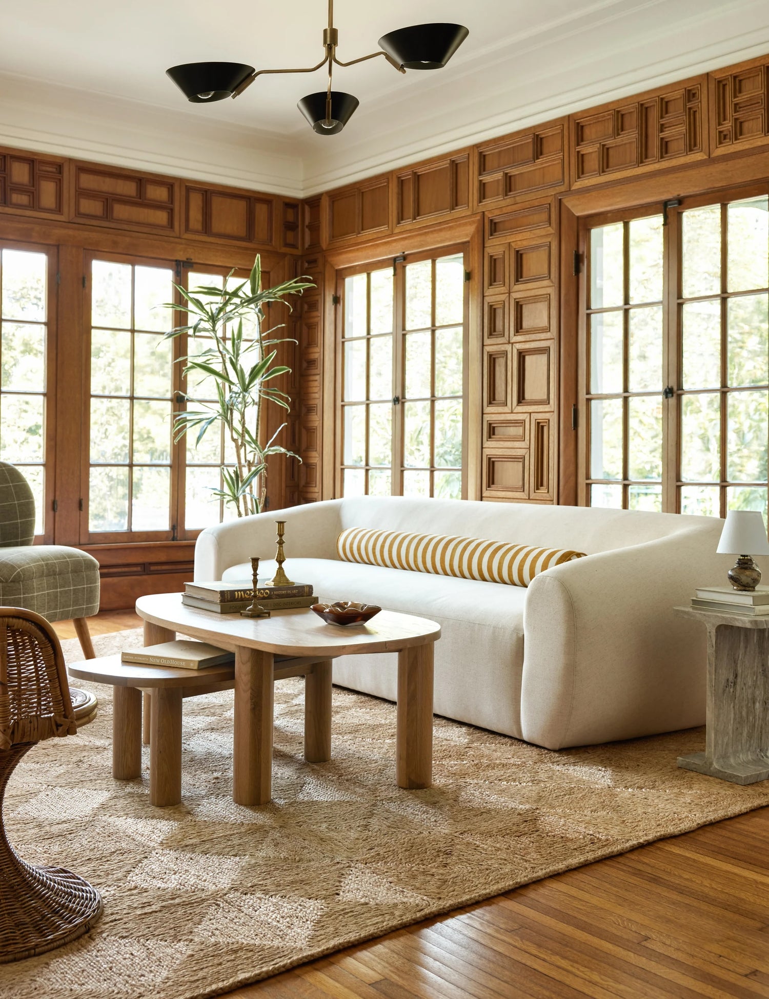 A stylish living room with wood-paneled walls and large, multi-paned windows. A white sofa with a striped bolster pillow sits on a patterned rug. A modern coffee table and chairs are arranged around it, and a tall plant is placed near the windows.