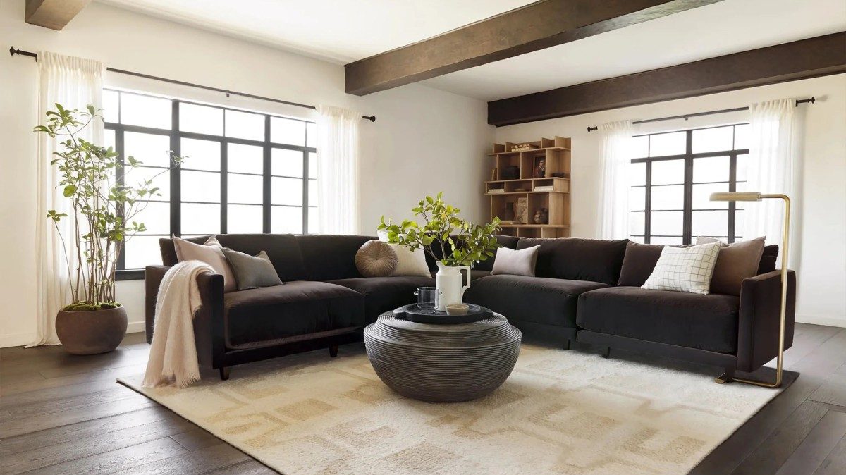 A modern living room with large windows framed by white curtains, dark wood beams on the ceiling, and a dark sectional sofa adorned with neutral pillows. A round black coffee table sits on a light patterned rug. Plants and a wooden bookshelf add decor elements.