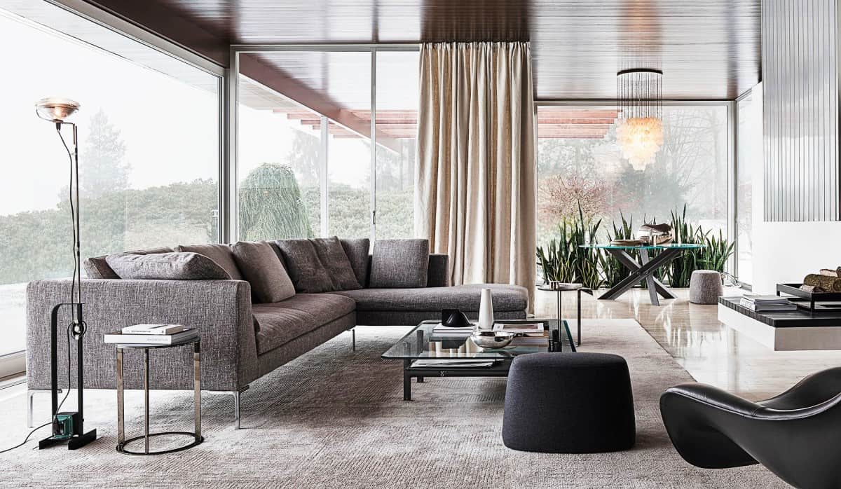 A modern living room with floor-to-ceiling windows. The space features a grey sectional sofa, glass coffee table, black ottoman, sleek black lounge chair, and a standing lamp. There is a tall plant next to the window, cream curtains, and a round dining table in the background.