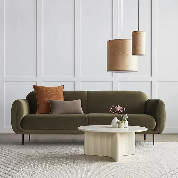 A modern living room featuring a green velvet sofa with brown and gray cushions. Two wooden pendant lights hang overhead. The white paneled wall adds to the minimalistic decor. In front of the sofa is a round, ribbed coffee table with small plants and a book on top.