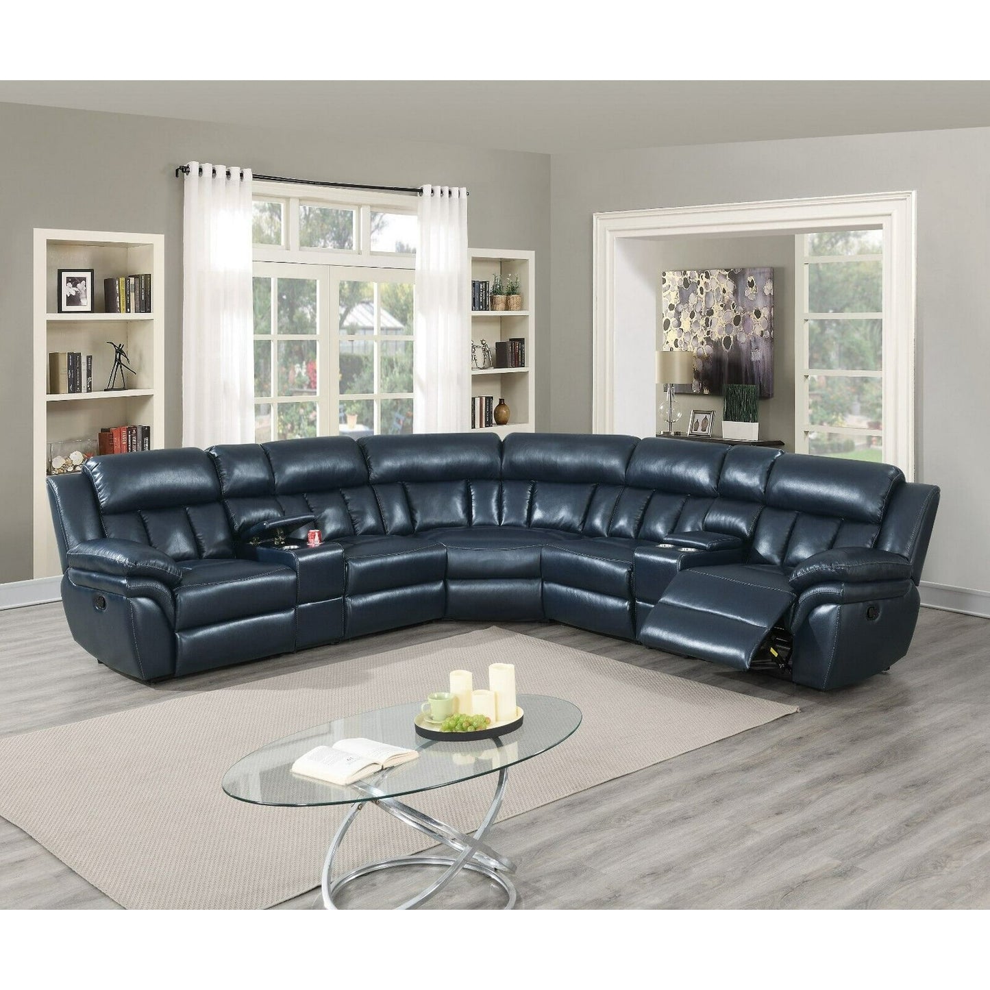 The spacious living room boasts a Luxurious Manual Motion Sectional Sofa in dark blue, arranged in a semi-circle. The space includes light-colored walls, a modern glass coffee table, and bookshelves. Large windows flood the room with natural light, accentuating the decor.