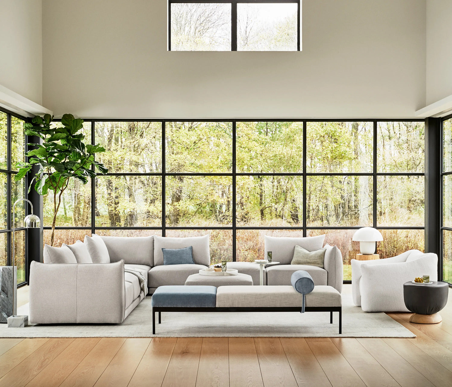 A modern living room with large floor-to-ceiling windows overlooking a wooded area. The space features light gray sofas, a white armchair, a cushioned bench, a round side table, a potted plant, and stylish decor pieces. There is a light-colored wooden floor.