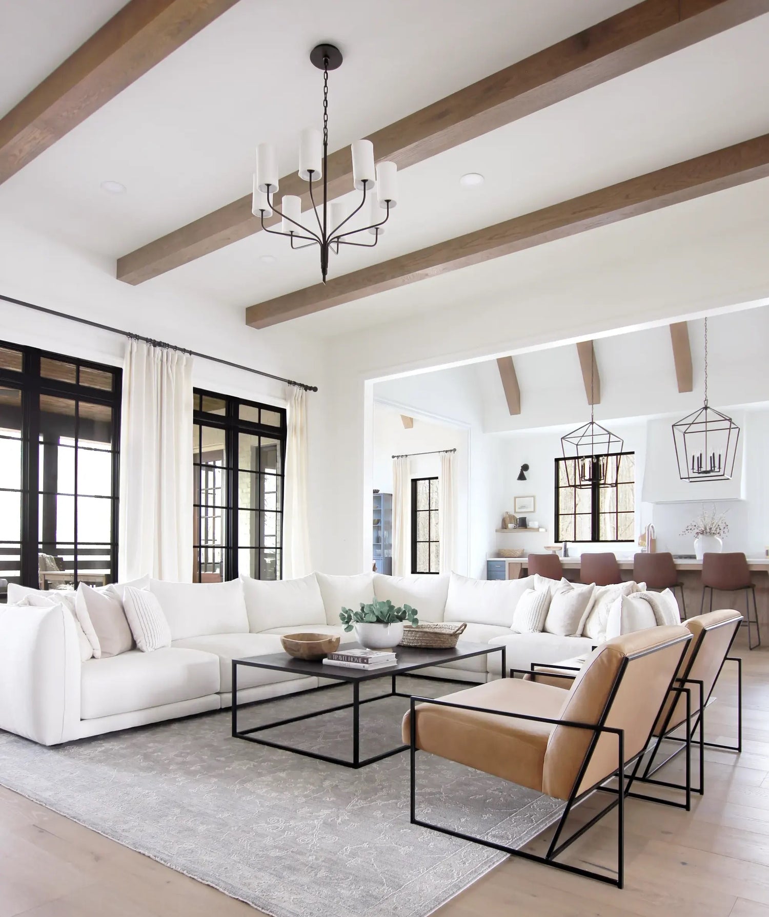 A modern, airy living room with white walls and wooden ceiling beams. It features a large white sectional sofa and two tan leather armchairs around a black metal coffee table. In the background, there's a dining area with a table and chairs beneath geometric light fixtures.