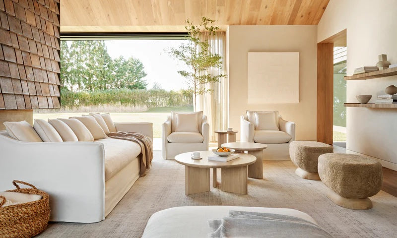 A modern living room with a white sofa, two matching armchairs, and two upholstered stools. A round coffee table holds decor items. Wooden shelves with ceramics adorn the wall. Large windows provide natural light and a view of greenery outside. Neutral tones dominate the space.