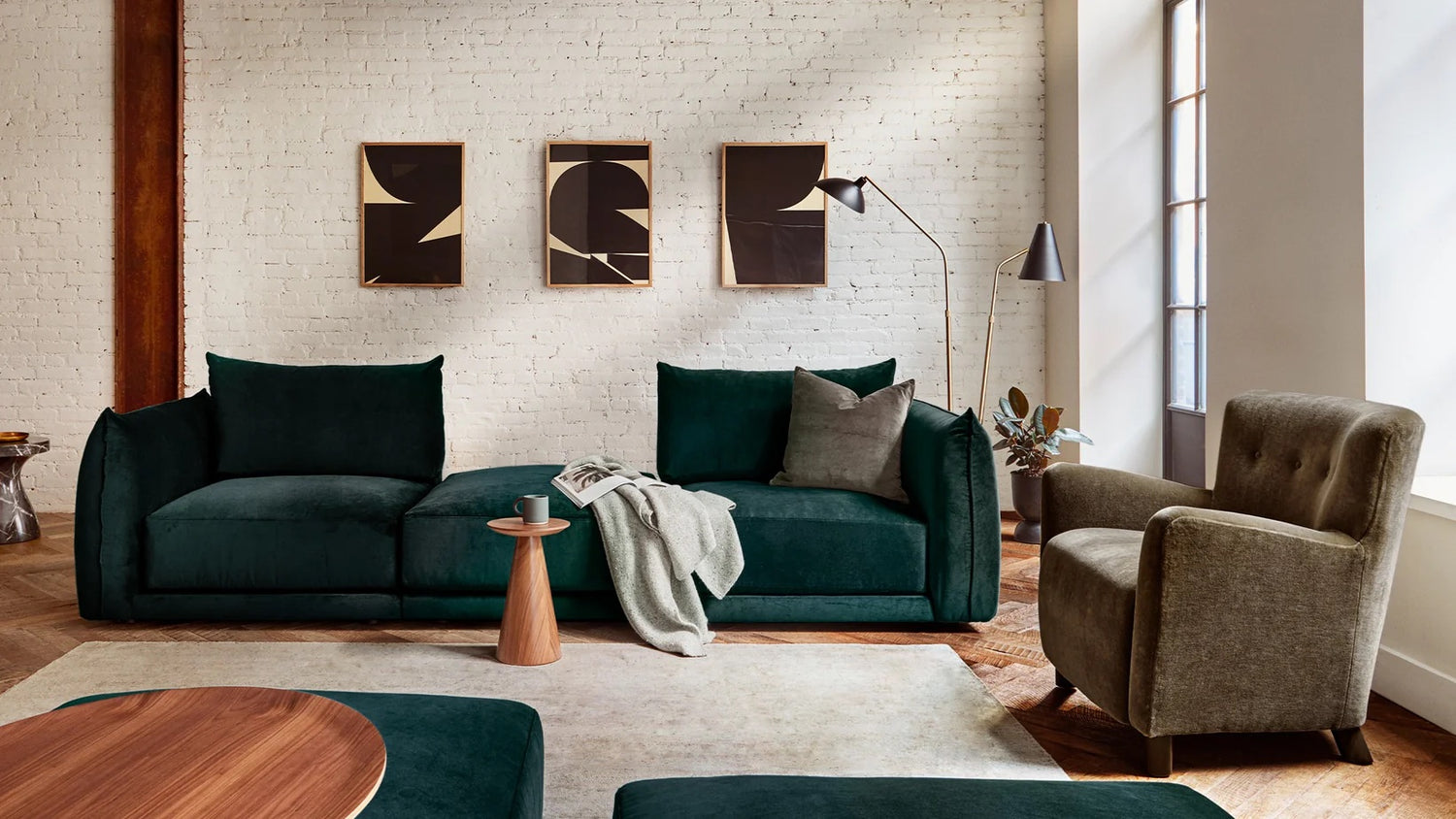 A modern living room with a dark green sofa, a gray armchair, a wooden side table with a magazine and a blanket on the sofa. Three abstract art pieces hang on a white brick wall, and a tall floor lamp stands next to the sofa. Sunlight streams through a window.