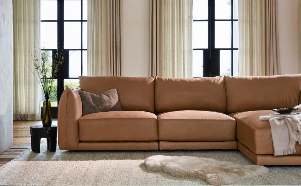 A modern living room features a tan sectional sofa with a grey pillow and a light throw blanket. Behind the sofa are large windows with light beige curtains. A small black side table with a vase holding dried plants stands to the left, and a soft rug covers the floor.
