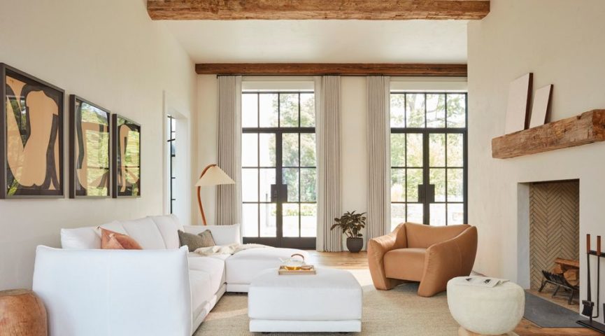 A cozy living room with a white couch, beige armchair, and a small ottoman. Large windows with light curtains allow natural light. The room features wooden ceiling beams, a fireplace with a wooden mantel, abstract art on the walls, and a floor lamp.