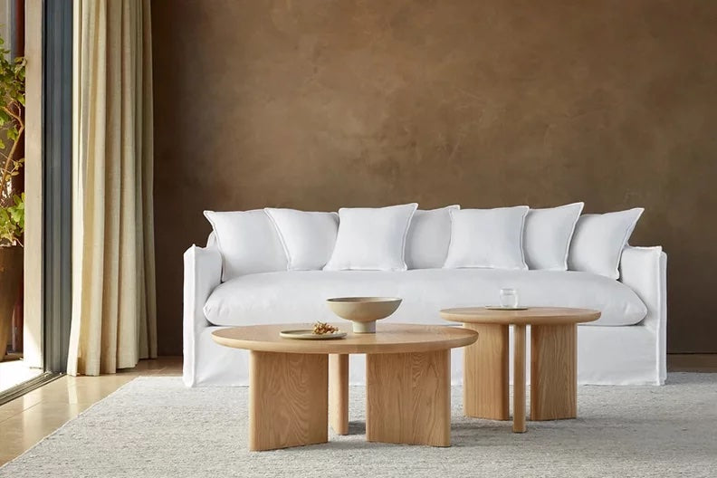 A modern living room features a white sofa with multiple white pillows, placed against a brown wall. In front of the sofa are two round wooden coffee tables, each holding decor items. The room has light curtains partially drawn open, letting in natural light.