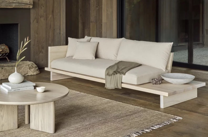 A minimalist living room with a low, light wood sofa featuring cream cushions and a gray throw blanket. A round wooden coffee table with decorative items, including a white bowl and vase, sits on a textured beige rug. Dark wooden walls and large windows frame the room.