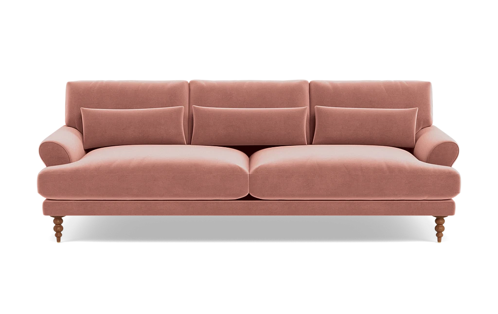 The Maxwell Custom Sofa is a plush, pink velvet piece featuring three back cushions and four cylindrical accent pillows. It showcases rolled arms and is supported by turned wooden legs, offering a blend of classic and contemporary design.