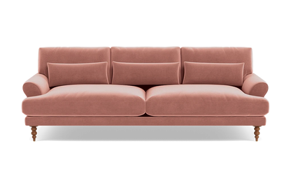 The Maxwell Custom Sofa is a plush, pink velvet piece featuring three back cushions and four cylindrical accent pillows. It showcases rolled arms and is supported by turned wooden legs, offering a blend of classic and contemporary design.