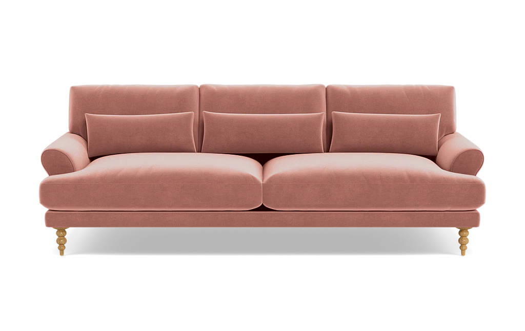 Introducing the Maxwell Custom Sofa: a charming pink velvet piece featuring three seat cushions and coordinating back cushions. It boasts elegantly rounded armrests and stands on four wooden legs adorned with intricate carvings. Enhancing its comfort are two rectangular throw pillows on the seat, all set against a simple black backdrop.