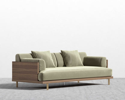 The Maria Sofa, with its light green cushions and wooden armrests, is placed on a light gray floor against a plain white wall. Its sleek design includes metal legs, giving it a minimalist and contemporary appearance.