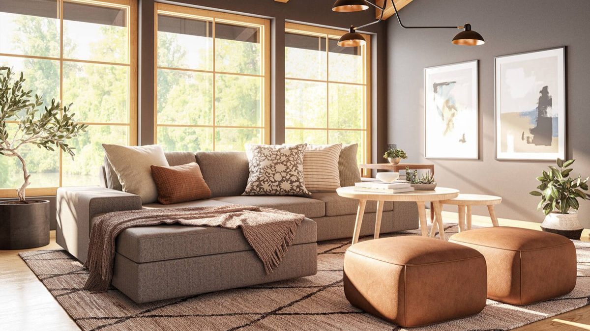 A cozy living room with a large gray sectional sofa adorned with patterned throw pillows and a blanket. A wooden coffee table, two brown ottomans, and framed art on the dark walls enhance the space. Large windows and indoor plants add brightness and warmth.