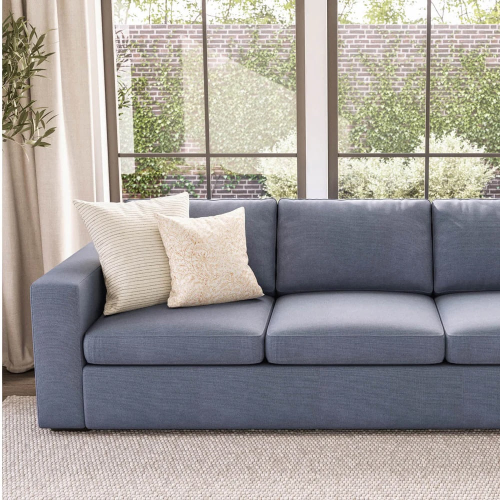 A modern living room features a blue sofa with clean lines. Two patterned throw pillows are placed on the left side of the sofa. Large windows in the background offer a view of lush greenery and a brick wall. Light curtains frame the windows.
