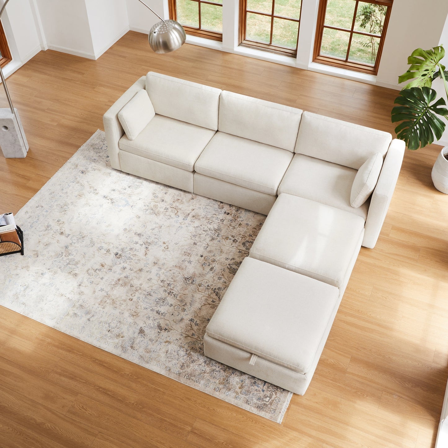 A top view of a modern living room showcases the Mia Oversized Modular Sectional 5-Piece Fabric Sofa Set in a light cream L-shape resting on a textured beige area rug. The room is adorned with wooden flooring, large windows, a vibrant green potted plant, and a contemporary floor lamp.