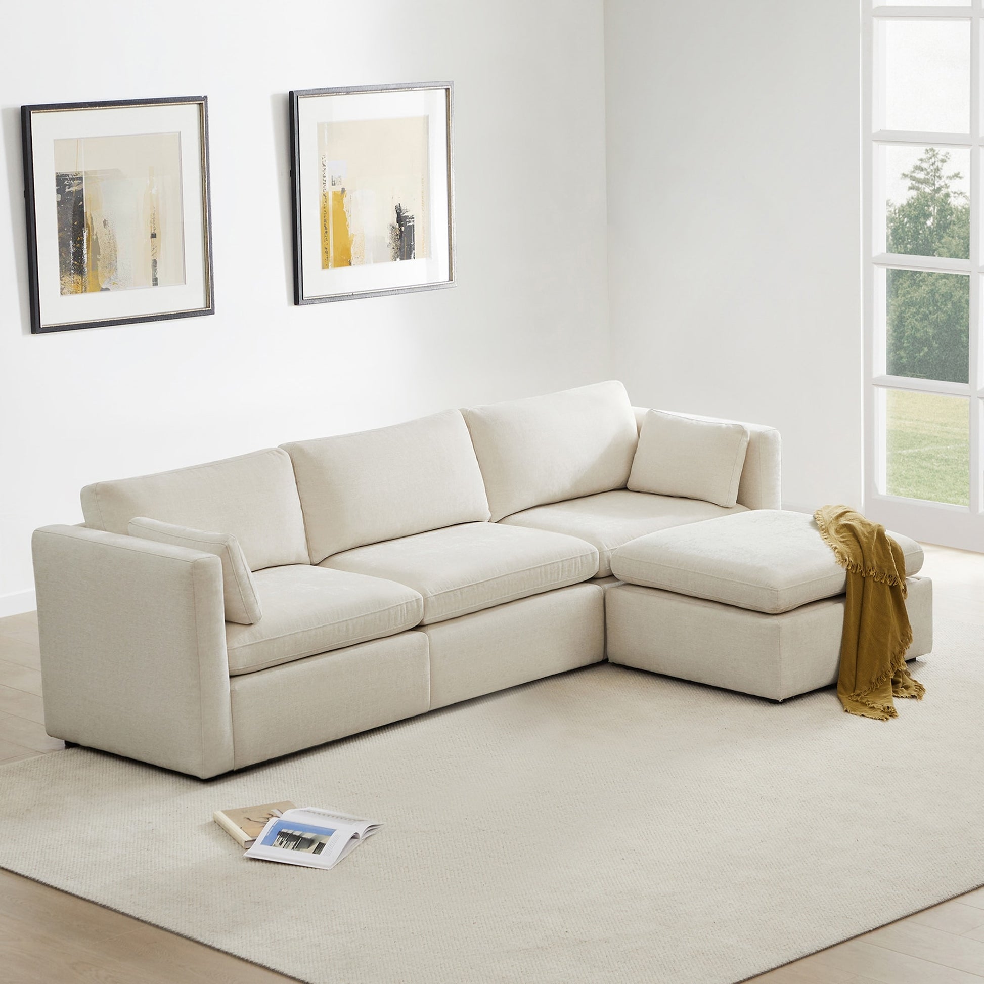 In a bright room, the Mia Oversized Modular Sectional 5-Piece Fabric Sofa Set, featuring a spacious L-shaped design and plush cushions in beige, rests on a light area rug. Two abstract art pieces adorn the wall, while a yellow throw blanket is casually draped over the sofa. A large window offers views of lush greenery outside.