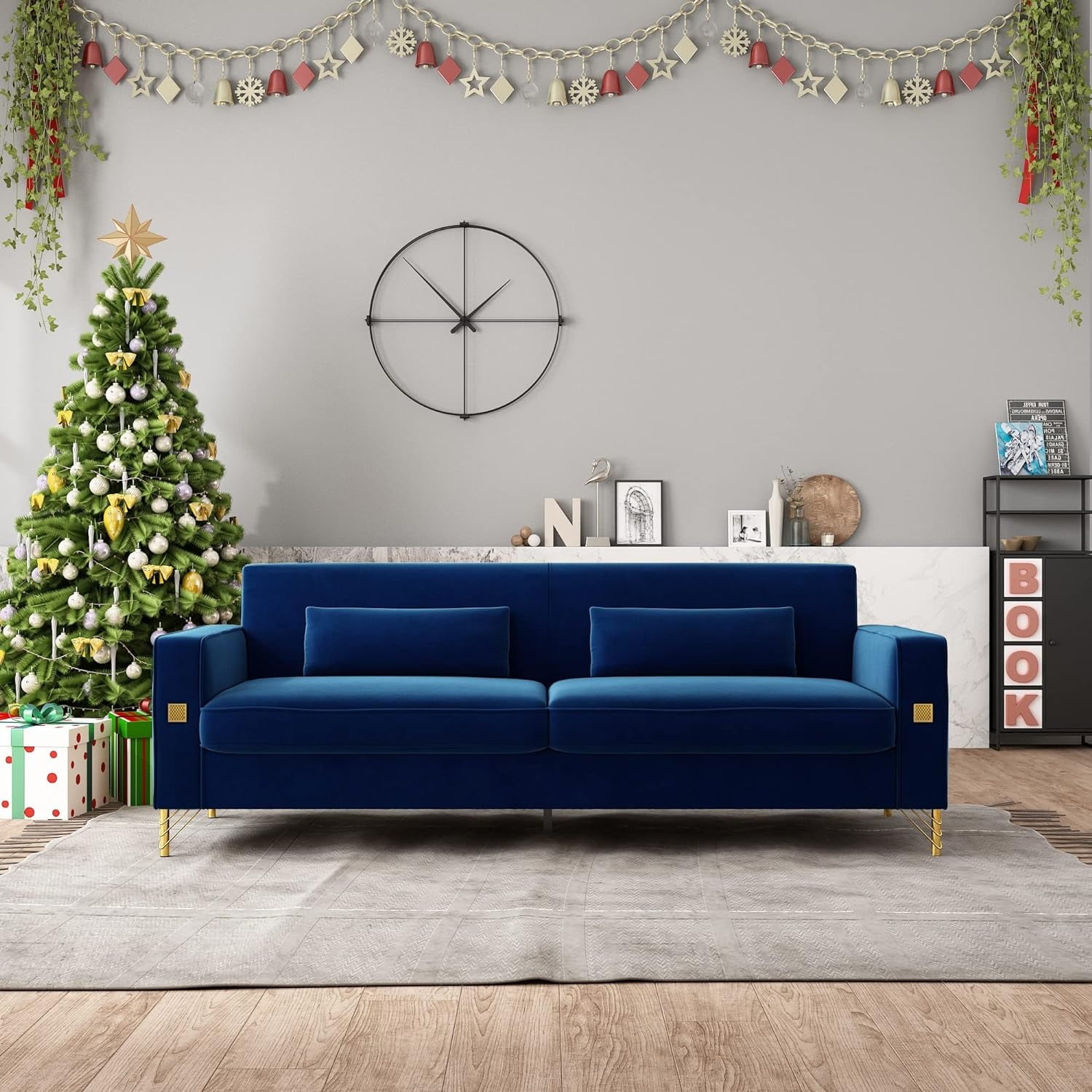 A modern living room features the Mid Century Modern Sofa Couch in a rich blue velvet upholstered on a gray rug. To the left, a beautifully decorated Christmas tree is surrounded by presents. Above the sofa hangs a large wall clock, while shelves filled with books and decor adorn the right side. Garland and plants enhance the festive atmosphere.