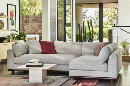 The room showcases a contemporary design with the Miller Sectional in light gray, embellished with red and striped cushions. It features a marble coffee table topped with books, while large windows reveal a view of lush greenery outside. Completing the aesthetic is a colorful rug.