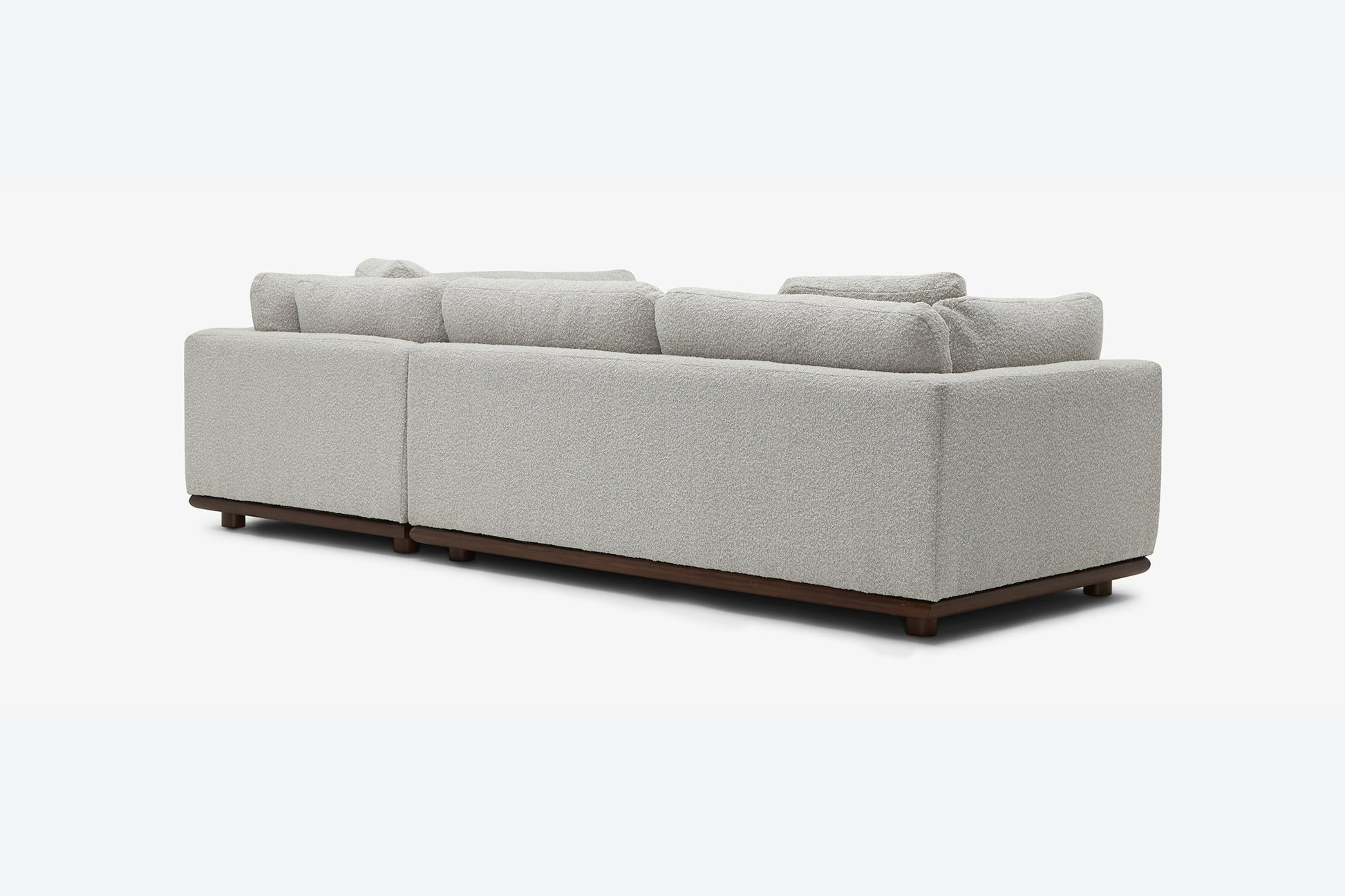 The Miller Sectional, a light gray modern sofa with a textured fabric and wooden base, is displayed from the back view. It features large, plush cushions and is supported by small, dark wooden legs.