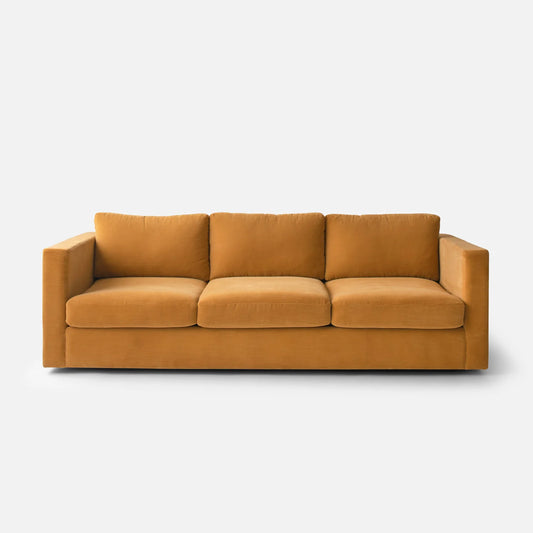 The Milo Sofa, a mustard-yellow three-seater with a minimalist design, is displayed against a plain white background. It features straight arms along with three cushioned seats and backs, offering a modern and cozy appearance.