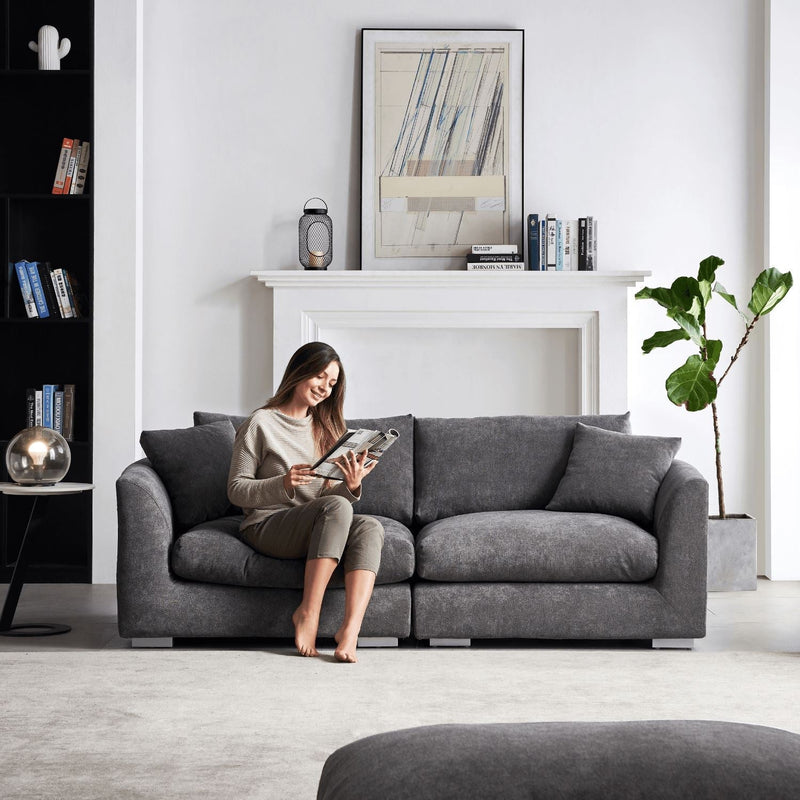 A woman relaxes on a Feathers 3-Seater Sofa in a contemporary living room, engrossed in a magazine. To the left stands a bookshelf, while to the right is a sizable plant, and above the white fireplace hangs framed artwork. The room is brightly lit and decorated with stylish flair.