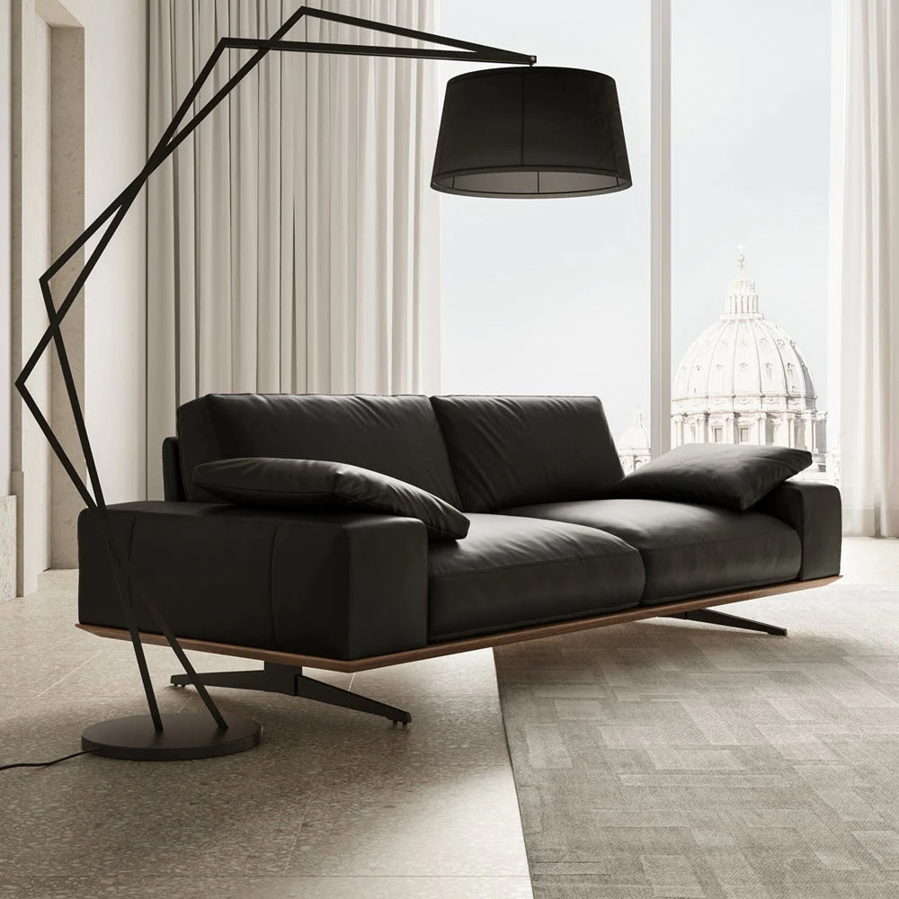 A modern living room with a sleek black leather sofa, two dark cushions, and a unique angular floor lamp with a large black shade. The room has large windows with white curtains, letting in natural light and offering a view of a dome structure outside.