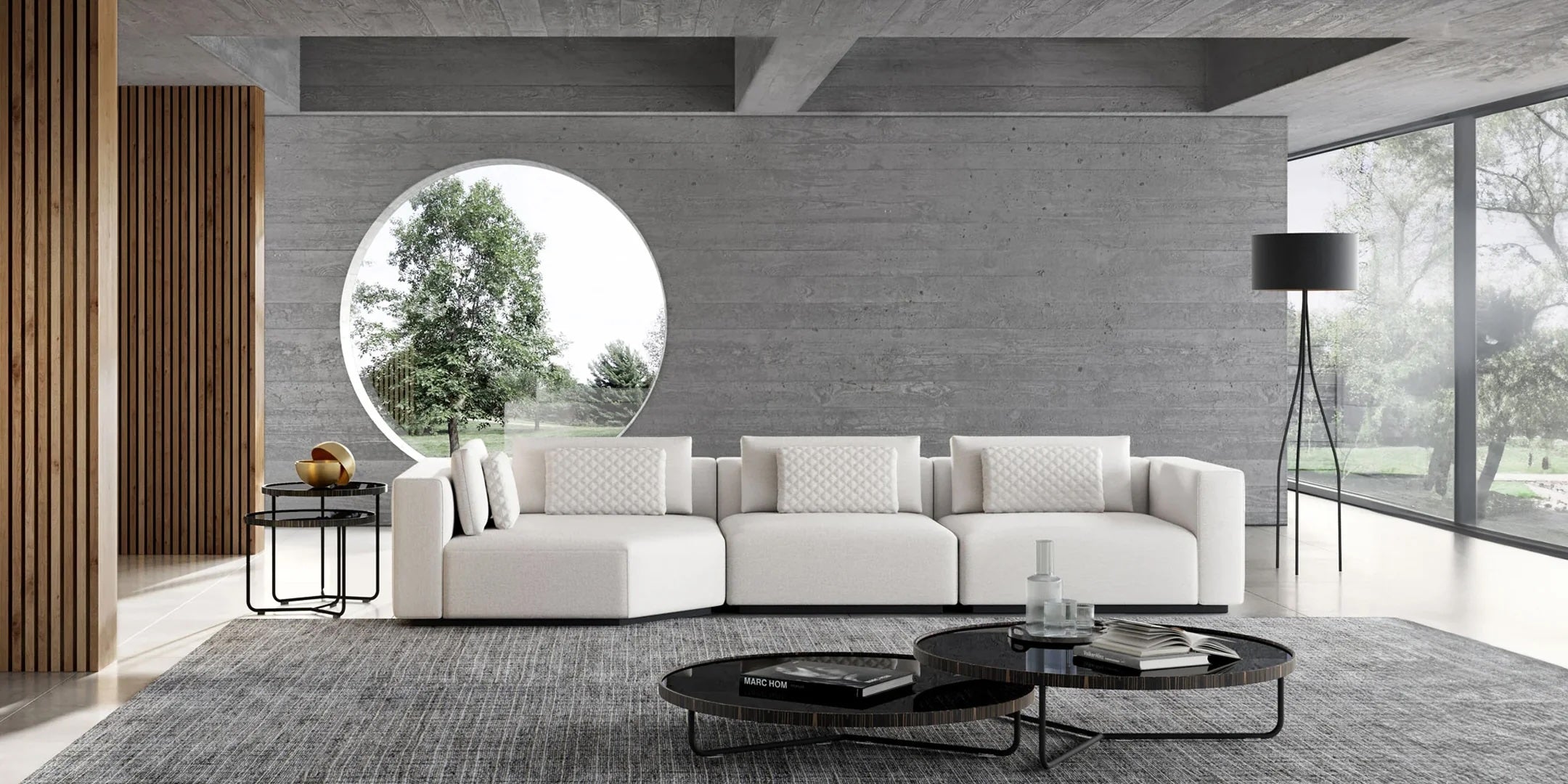 A modern living room with a minimalist design features a large white sectional sofa and a pair of round black coffee tables on a gray rug. A round window, floor lamp, and a side table with decorative items enhance the room's contemporary decor.