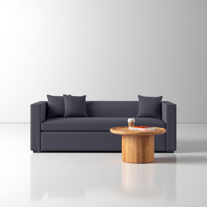 A minimalist room showcasing the Mono 81" Sleeper Sofa in dark gray, accompanied by two matching cushions. Positioned in front of the sofa is a round wooden coffee table holding a stack of books and a paper cup, all framed by a light gray wall and floor.