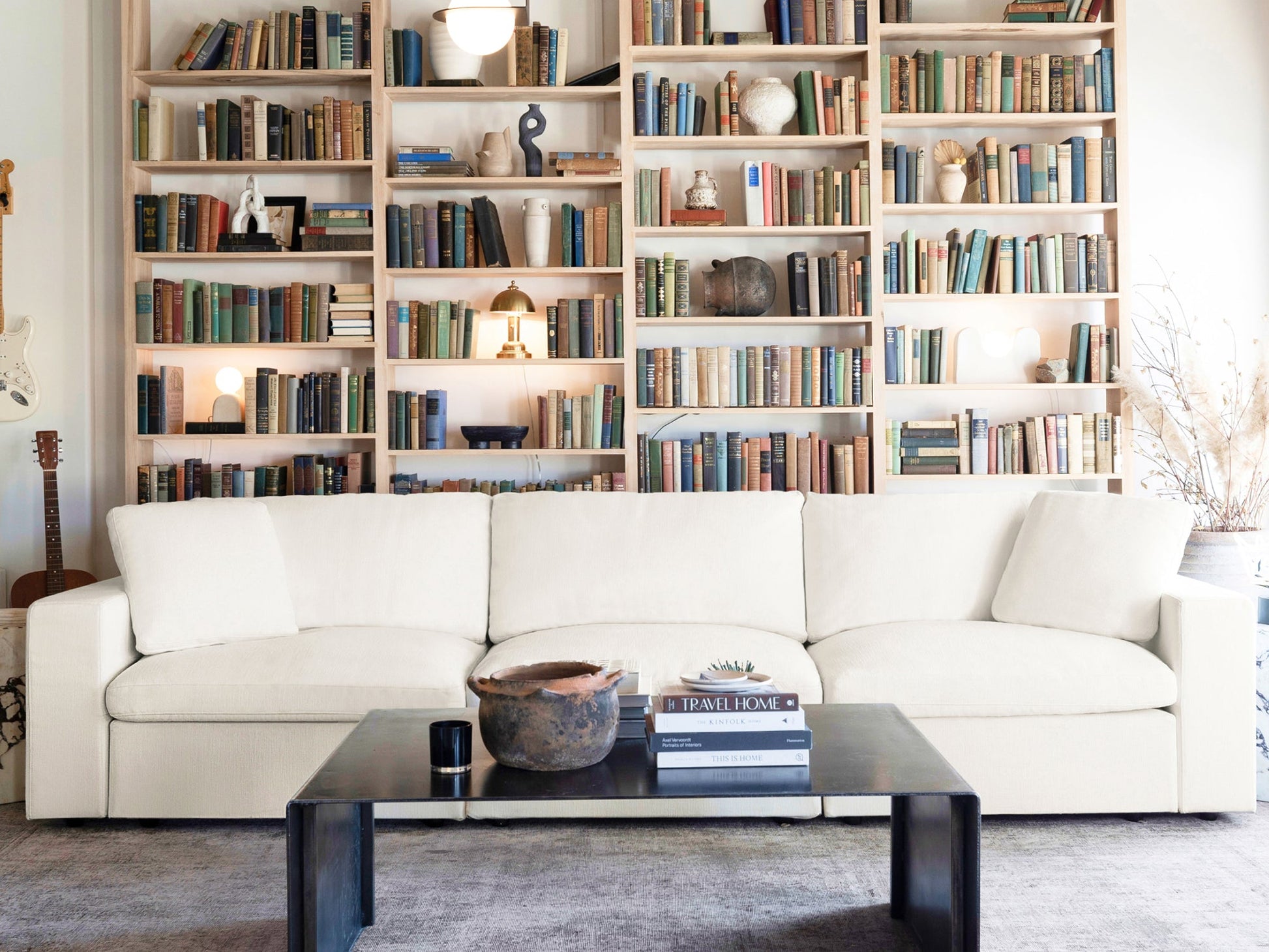 In a cozy living room, a 2-Seat Modular Loveseat sits in front of a wall lined with tall bookshelves filled with books and decorative items. A black coffee table displays a few stacked books and a clay pot, while soft lighting enhances the warm and inviting atmosphere.