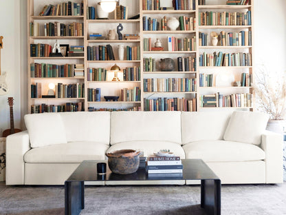 In a cozy living room, a 2-Seat Modular Loveseat sits in front of a wall lined with tall bookshelves filled with books and decorative items. A black coffee table displays a few stacked books and a clay pot, while soft lighting enhances the warm and inviting atmosphere.