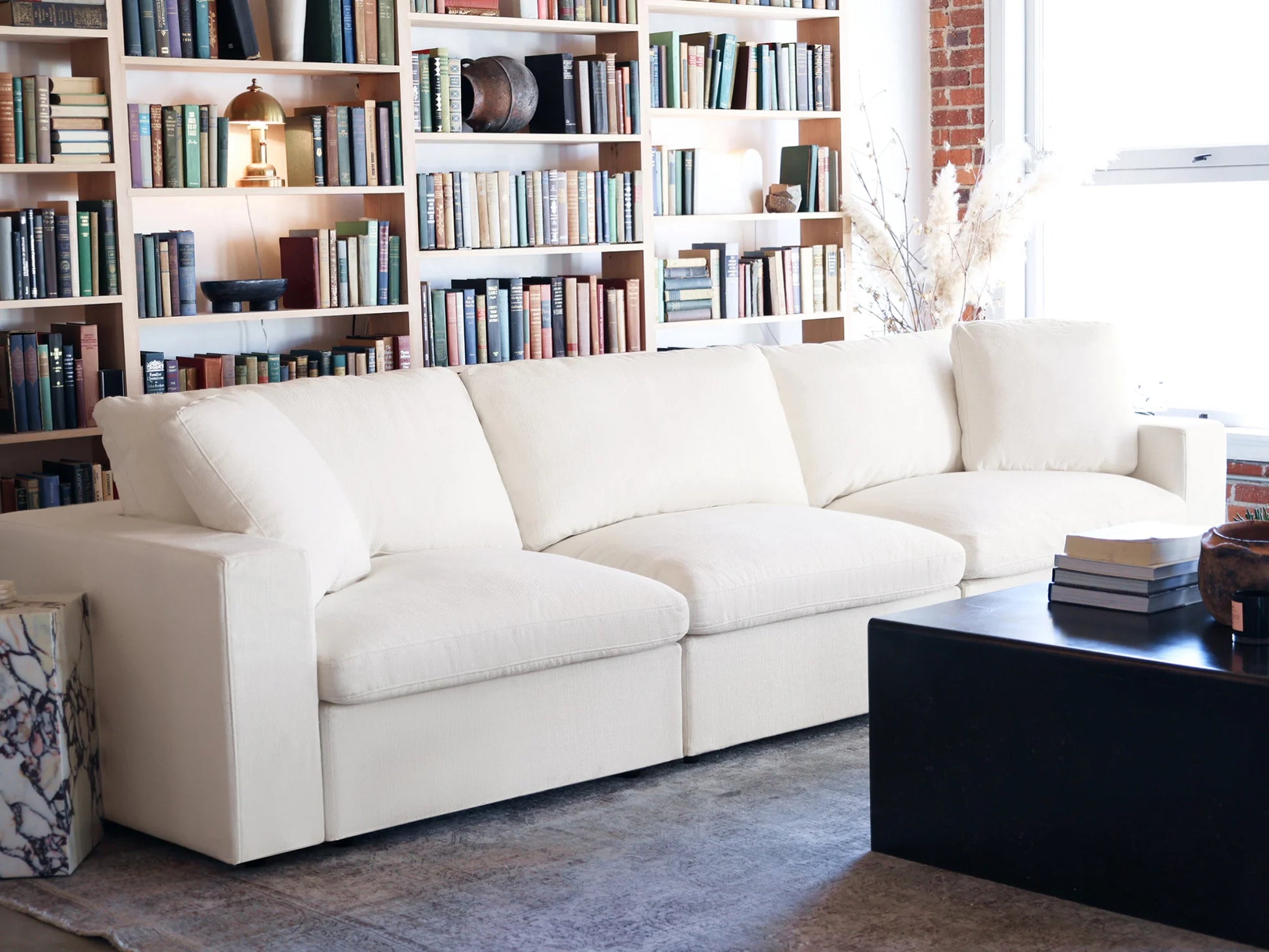 A cozy living room features a spacious 4-Seat Modular Chaise Sectional with Ottoman in white, positioned in front of a wall adorned with bookshelves filled with various books and decor. A dark coffee table, neatly stacked with books, rests on a patterned rug, while natural light pours in through the window.