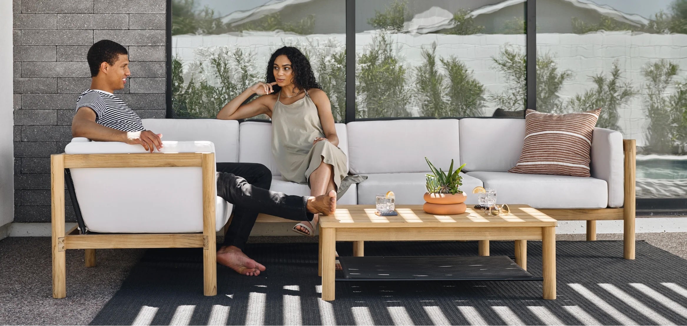 A man and a woman sit on a white outdoor sofa set with wooden frames. The man is smiling, and the woman looks thoughtful. A wooden coffee table adorned with plants and decorative items is in front of them. They are on a patio with glass windows and potted plants.