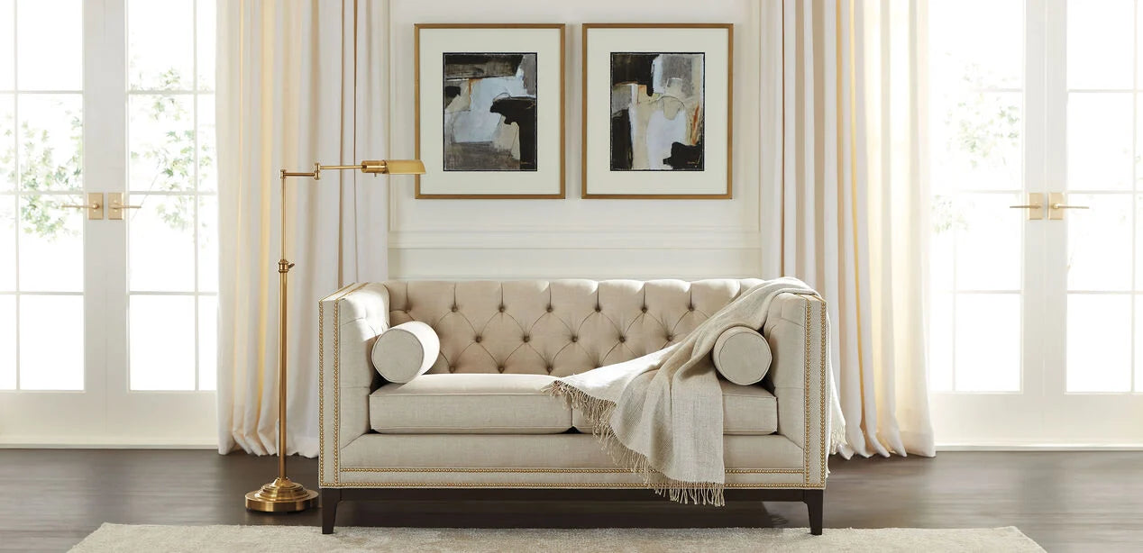 The Anderson Loveseat in beige with a throw blanket is placed against a backdrop of floor-to-ceiling curtains. Two abstract art pieces adorn the wall above the loveseat. To the left stands a tall, gold floor lamp, and dark wood flooring completes the room’s aesthetic.