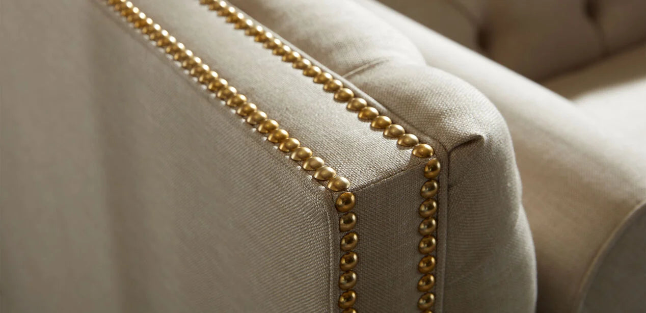 Close-up of the Anderson Loveseat's beige upholstered armrest, featuring a trim of decorative brass nailhead accents along the edge, adding a touch of elegance to the furniture.